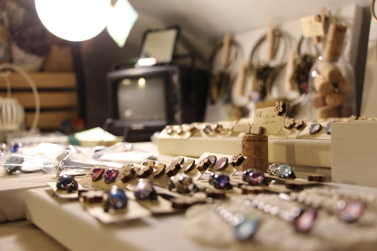 A collection of artisan jewelry displayed on a table with various pieces, including rings and necklaces, featuring colorful stones. Soft focus in the background highlights a vintage television and some decorative items, such as corks and glass containers. Warm lighting illuminates the scene, creating a cozy and inviting atmosphere.