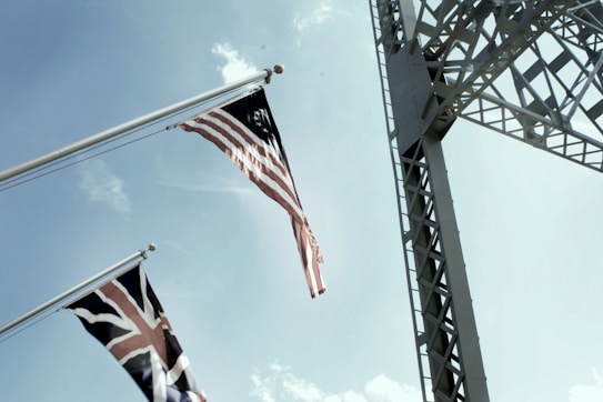 Two flags, one with the American flag design and another with the British Union Jack, are waving in the wind against a clear blue sky. A large metal structure with intricate latticework extends vertically through the right side of the image.