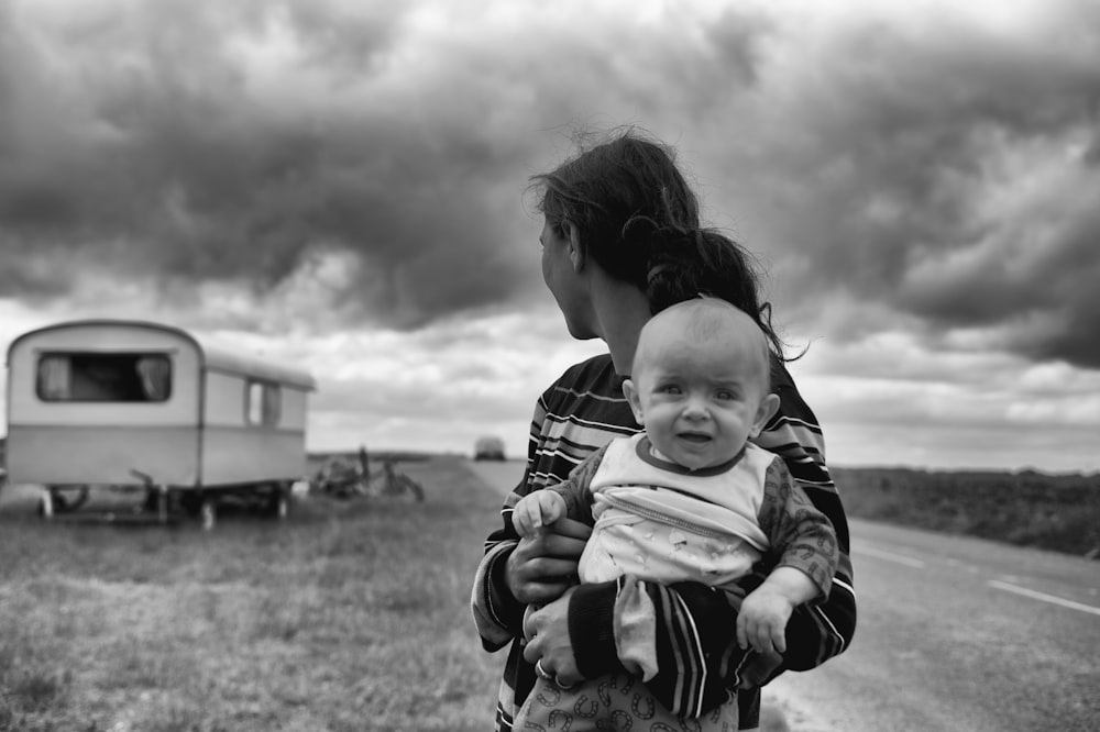 gray scale photography of woman carrying baby looking at camper trailer