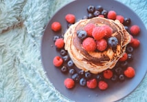 A stack of pancakes topped with fresh raspberries and blueberries is arranged on a dark plate. The plate is surrounded by a soft, textured light blue background, adding a cozy feel.