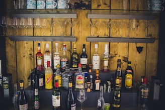A variety of liquor bottles are arranged on shelves against a wooden wall. Above the bottles, there are several wine and cocktail glasses neatly organized. The setup includes well-known brands, and the lighting gives a rustic ambiance.
