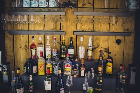 A variety of liquor bottles are arranged on shelves against a wooden wall. Above the bottles, there are several wine and cocktail glasses neatly organized. The setup includes well-known brands, and the lighting gives a rustic ambiance.