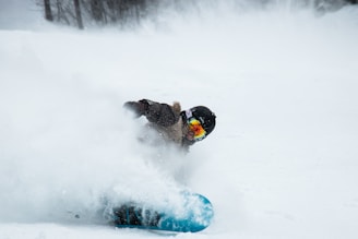 A person is snowboarding on a snowy slope, kicking up snow. They are wearing a helmet and goggles with reflective lenses, and a winter jacket. The snowboarder is leaning sharply into the turn, with snow flying around them.