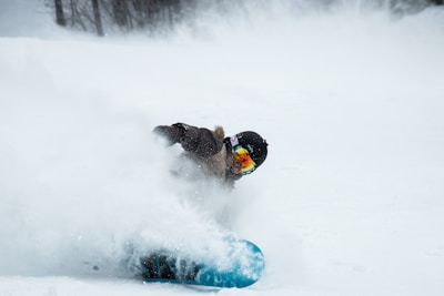 A person is snowboarding on a snowy slope, kicking up snow. They are wearing a helmet and goggles with reflective lenses, and a winter jacket. The snowboarder is leaning sharply into the turn, with snow flying around them.