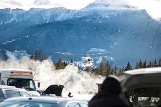 A helicopter hovers above the snowy landscape with mountains in the background. A group of vehicles, including a bus labeled 'Resort Express,' are parked in the foreground. Snow is being blown around, creating a misty effect.