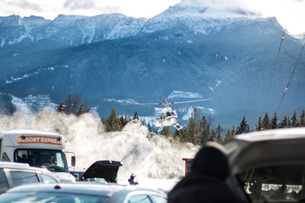 A helicopter hovers above the snowy landscape with mountains in the background. A group of vehicles, including a bus labeled 'Resort Express,' are parked in the foreground. Snow is being blown around, creating a misty effect.