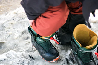 A skier adjusting bindings on sturdy boots beside a snowy mountain slope.