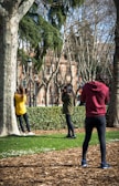 Three people appear to be engaged in a photography session in a park. A woman in a yellow top leans against a tree while another person, wearing a green jacket, takes her photo with a camera. A third person, in a red hoodie, is also taking photographs. The background features trees with bare branches and a brick building. The ground is covered with wood chips and a grassy area with small white flowers.