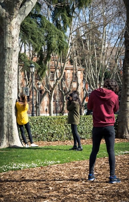 Three people appear to be engaged in a photography session in a park. A woman in a yellow top leans against a tree while another person, wearing a green jacket, takes her photo with a camera. A third person, in a red hoodie, is also taking photographs. The background features trees with bare branches and a brick building. The ground is covered with wood chips and a grassy area with small white flowers.