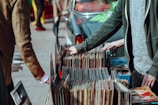 A street market scene with people browsing through a collection of vintage vinyl records. The records are neatly organized in boxes on a table set up against a car. Two individuals are visible, one wearing a green jacket and another in a brown coat. Their attention is focused on selecting records, suggesting an interest in music.