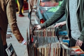 A street market scene with people browsing through a collection of vintage vinyl records. The records are neatly organized in boxes on a table set up against a car. Two individuals are visible, one wearing a green jacket and another in a brown coat. Their attention is focused on selecting records, suggesting an interest in music.