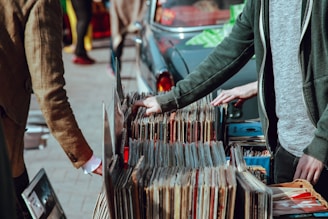 A street market scene with people browsing through a collection of vintage vinyl records. The records are neatly organized in boxes on a table set up against a car. Two individuals are visible, one wearing a green jacket and another in a brown coat. Their attention is focused on selecting records, suggesting an interest in music.