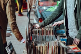 A street market scene with people browsing through a collection of vintage vinyl records. The records are neatly organized in boxes on a table set up against a car. Two individuals are visible, one wearing a green jacket and another in a brown coat. Their attention is focused on selecting records, suggesting an interest in music.