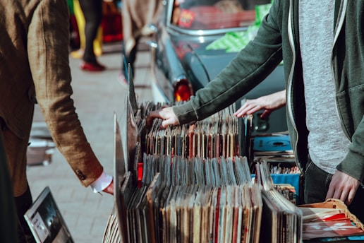 A street market scene with people browsing through a collection of vintage vinyl records. The records are neatly organized in boxes on a table set up against a car. Two individuals are visible, one wearing a green jacket and another in a brown coat. Their attention is focused on selecting records, suggesting an interest in music.