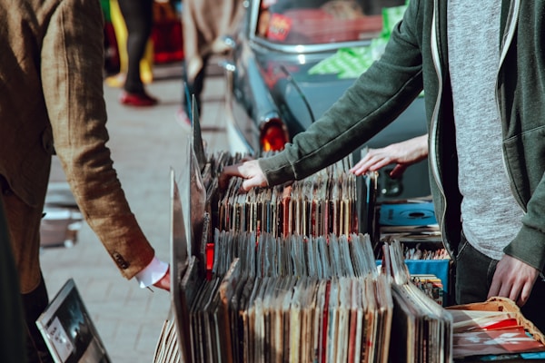 A street market scene with people browsing through a collection of vintage vinyl records. The records are neatly organized in boxes on a table set up against a car. Two individuals are visible, one wearing a green jacket and another in a brown coat. Their attention is focused on selecting records, suggesting an interest in music.