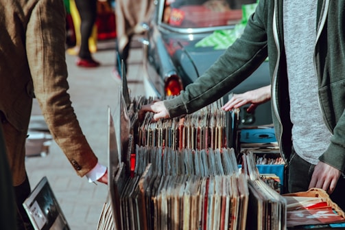 A street market scene with people browsing through a collection of vintage vinyl records. The records are neatly organized in boxes on a table set up against a car. Two individuals are visible, one wearing a green jacket and another in a brown coat. Their attention is focused on selecting records, suggesting an interest in music.