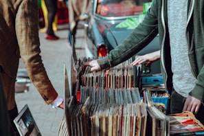 A street market scene with people browsing through a collection of vintage vinyl records. The records are neatly organized in boxes on a table set up against a car. Two individuals are visible, one wearing a green jacket and another in a brown coat. Their attention is focused on selecting records, suggesting an interest in music.