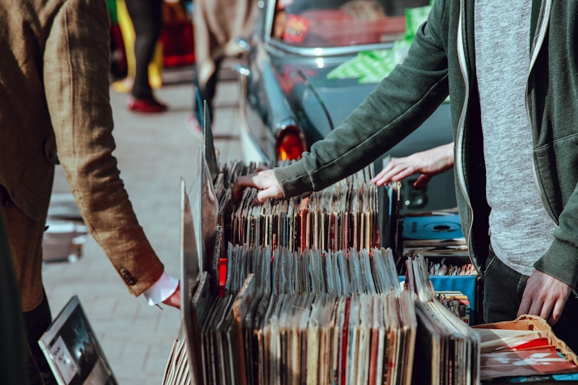 A street market scene with people browsing through a collection of vintage vinyl records. The records are neatly organized in boxes on a table set up against a car. Two individuals are visible, one wearing a green jacket and another in a brown coat. Their attention is focused on selecting records, suggesting an interest in music.