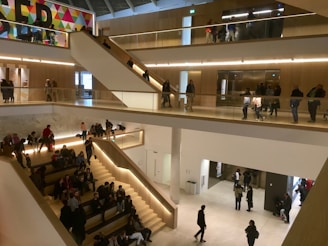 A multi-level indoor space with people walking and sitting. The architecture includes modern staircases, glass railings, and ample natural light. A colorful geometric mural can be seen on one wall.