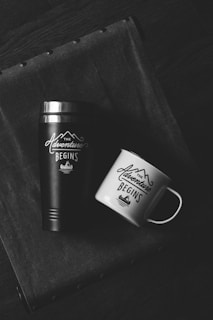 Two drinking containers are placed on a textured fabric surface, a black thermos and a white mug both labeled with 'The Adventure Begins' and a mountain graphic.