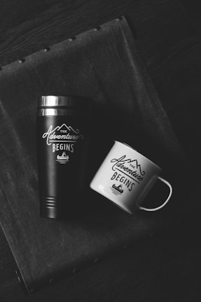 Two drinking containers are placed on a textured fabric surface, a black thermos and a white mug both labeled with 'The Adventure Begins' and a mountain graphic.