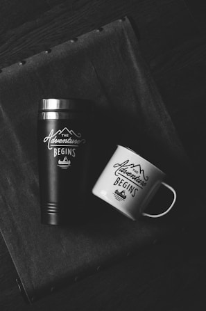 Two drinking containers are placed on a textured fabric surface, a black thermos and a white mug both labeled with 'The Adventure Begins' and a mountain graphic.