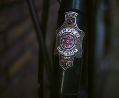 Close-up view of a vintage bicycle headbadge that reads 'Union Holland.' The badge is attached to a dark green bicycle frame and has a red gear-like design at its center, along with decorative elements.