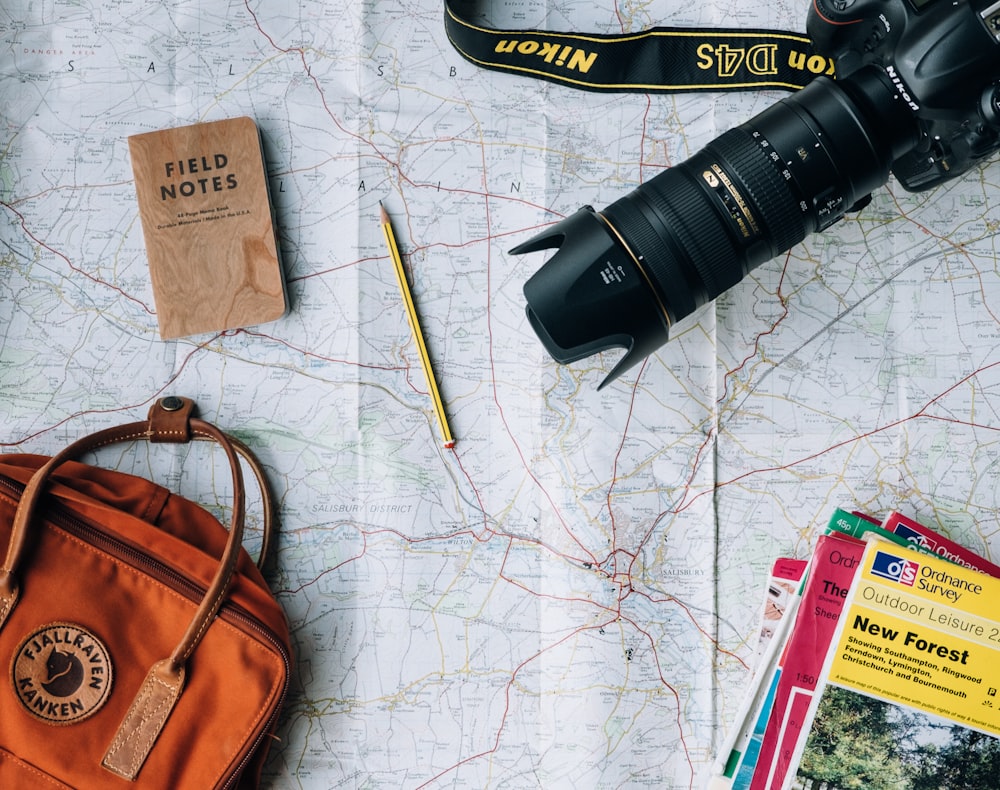 An overhead shot of a long-lens camera, a Field Notes Notebook, a pencil and a travel bag laid out on a map