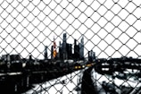 A chain-link fence in the foreground with a blurred city skyline in the background. The focus on the fence creates a sense of separation from the cityscape, which appears dark and shadowy against a bright sky.