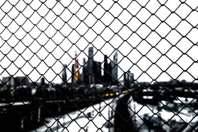A chain-link fence in the foreground with a blurred city skyline in the background. The focus on the fence creates a sense of separation from the cityscape, which appears dark and shadowy against a bright sky.