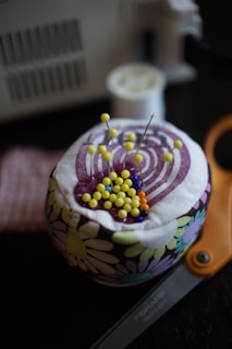 A pincushion covered with a floral fabric holds pins with colorful heads, predominantly yellow, with some blue and orange. Scissors labeled 'Fiskars' are partially visible in the foreground. A spool of thread and a sewing machine are blurred in the background.