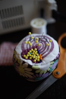 A pincushion covered with a floral fabric holds pins with colorful heads, predominantly yellow, with some blue and orange. Scissors labeled 'Fiskars' are partially visible in the foreground. A spool of thread and a sewing machine are blurred in the background.