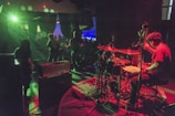 A band performing on stage with dynamic lighting and vibrant colors. The drummer is prominently visible, energetically playing, while other band members are playing guitars. The lights create a dramatic atmosphere with green and red hues. The audience is visible in the background.