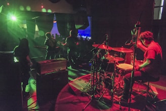 A band performing on stage with dynamic lighting and vibrant colors. The drummer is prominently visible, energetically playing, while other band members are playing guitars. The lights create a dramatic atmosphere with green and red hues. The audience is visible in the background.