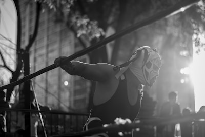 A masked wrestler in a lucha libre mask stands in a wrestling ring, leaning on the ropes. The image is in black and white, with sunlight filtering through the surrounding trees, creating a dramatic silhouette against an urban backdrop.