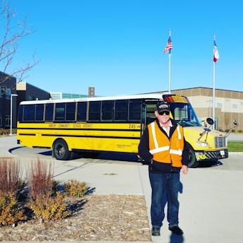 A bright yellow school bus with 'Ankeny Community School District' written on the side is parked on a pavement under a clear blue sky. In the foreground, a man wearing a high-visibility orange vest stands smiling. Flags are visible in the background, and the surrounding area includes a building and some landscaped plants.