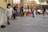 A young child in casual clothing and a backwards cap is running on a paved street beside a skateboard. The scene is lively with numerous people walking and socializing in the background. The architecture features a mix of colorful buildings and a stone column.