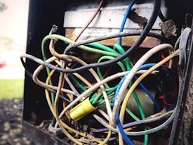 A chaotic tangle of multicolored electrical wires, including green, blue, black, and yellow, in what appears to be an electrical box. Some wires are frayed or damaged, and there is a yellow wire connector in the mix. The scene suggests neglect or disrepair.