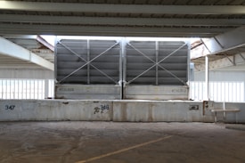 Large metallic cooling units are positioned under a steel-beamed structure in an industrial setting. The units have a grid design on their fronts and are elevated on a concrete platform marked with numbers 347 and 348. The scene is partially lit by natural light filtering through the wide windows behind them, and there are visible signs of rust on the beams.