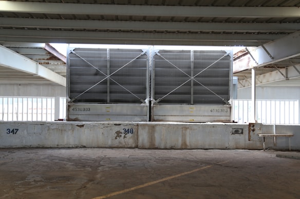 Large metallic cooling units are positioned under a steel-beamed structure in an industrial setting. The units have a grid design on their fronts and are elevated on a concrete platform marked with numbers 347 and 348. The scene is partially lit by natural light filtering through the wide windows behind them, and there are visible signs of rust on the beams.