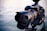 A professional DSLR camera is mounted on a tripod with a large lens extending from the body. The focus is on the camera and lens, with a blurred background of water.