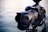 A professional DSLR camera is mounted on a tripod with a large lens extending from the body. The focus is on the camera and lens, with a blurred background of water.