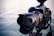 A professional DSLR camera is mounted on a tripod with a large lens extending from the body. The focus is on the camera and lens, with a blurred background of water.