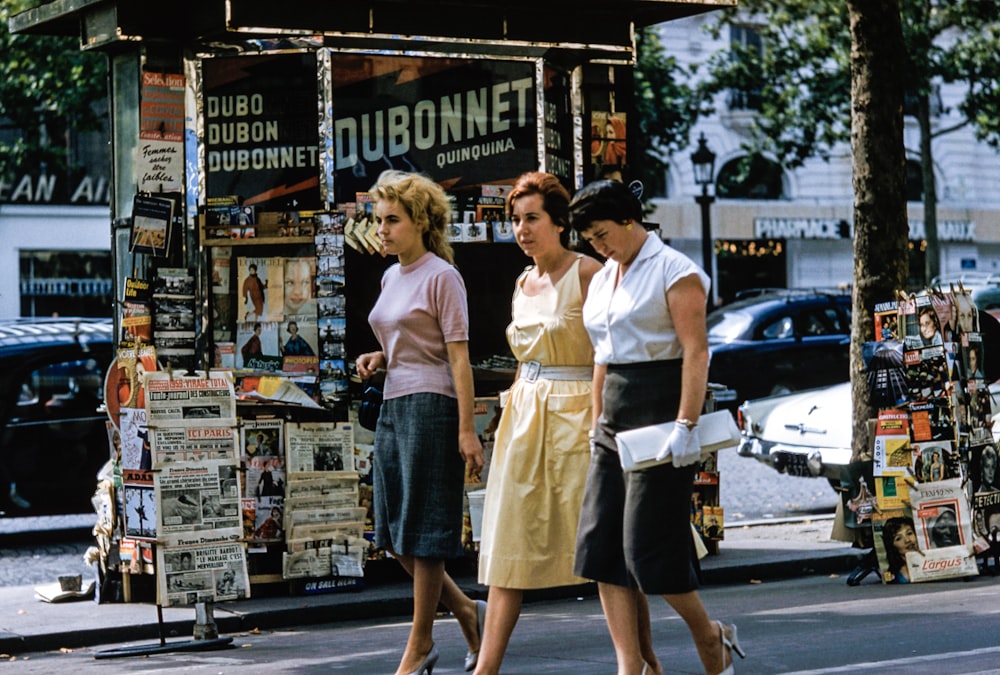 Three women in retro clothes walk on the sidewalk by a news stand