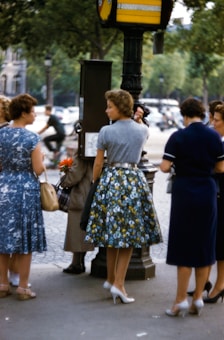 A group of women standing on a city sidewalk, engaging in conversation near a lamp post. They are dressed in mid-20th-century fashion, with dresses and skirts featuring floral patterns and solid colors. Some are holding handbags, and one woman is holding a flower. Trees and blurred cars are visible in the background, indicating an urban setting.