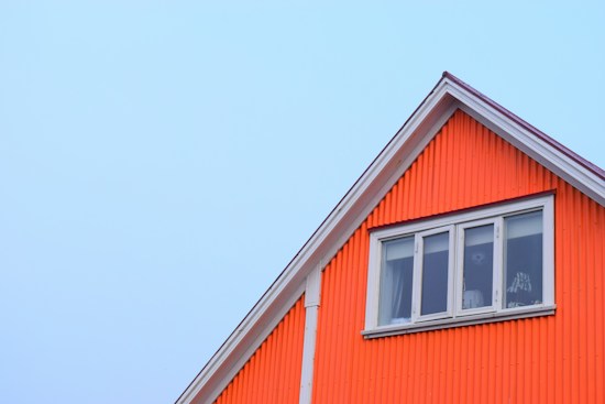 A vibrant orange house with a triangular roof section is pictured against a clear blue sky. The house exterior features vertical corrugations, and there is a set of white-framed windows allowing a glimpse inside.