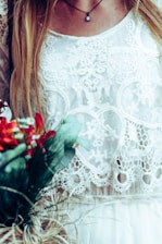 A detailed lace fabric with intricate patterns is featured, worn by a woman who also has long blonde hair cascading down. A delicate necklace with a small pendant hangs from her neck. The image includes a bouquet of vibrant flowers with green leaves in the lower left corner, adding a contrast to the soft textile patterns.