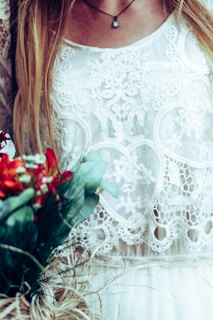 A detailed lace fabric with intricate patterns is featured, worn by a woman who also has long blonde hair cascading down. A delicate necklace with a small pendant hangs from her neck. The image includes a bouquet of vibrant flowers with green leaves in the lower left corner, adding a contrast to the soft textile patterns.