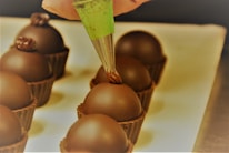 A hand holding a green piping bag applies a decorative chocolate topping onto round chocolate truffles placed in brown chocolate cups, arranged neatly on a light-colored surface.