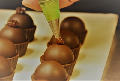 A hand holding a green piping bag applies a decorative chocolate topping onto round chocolate truffles placed in brown chocolate cups, arranged neatly on a light-colored surface.